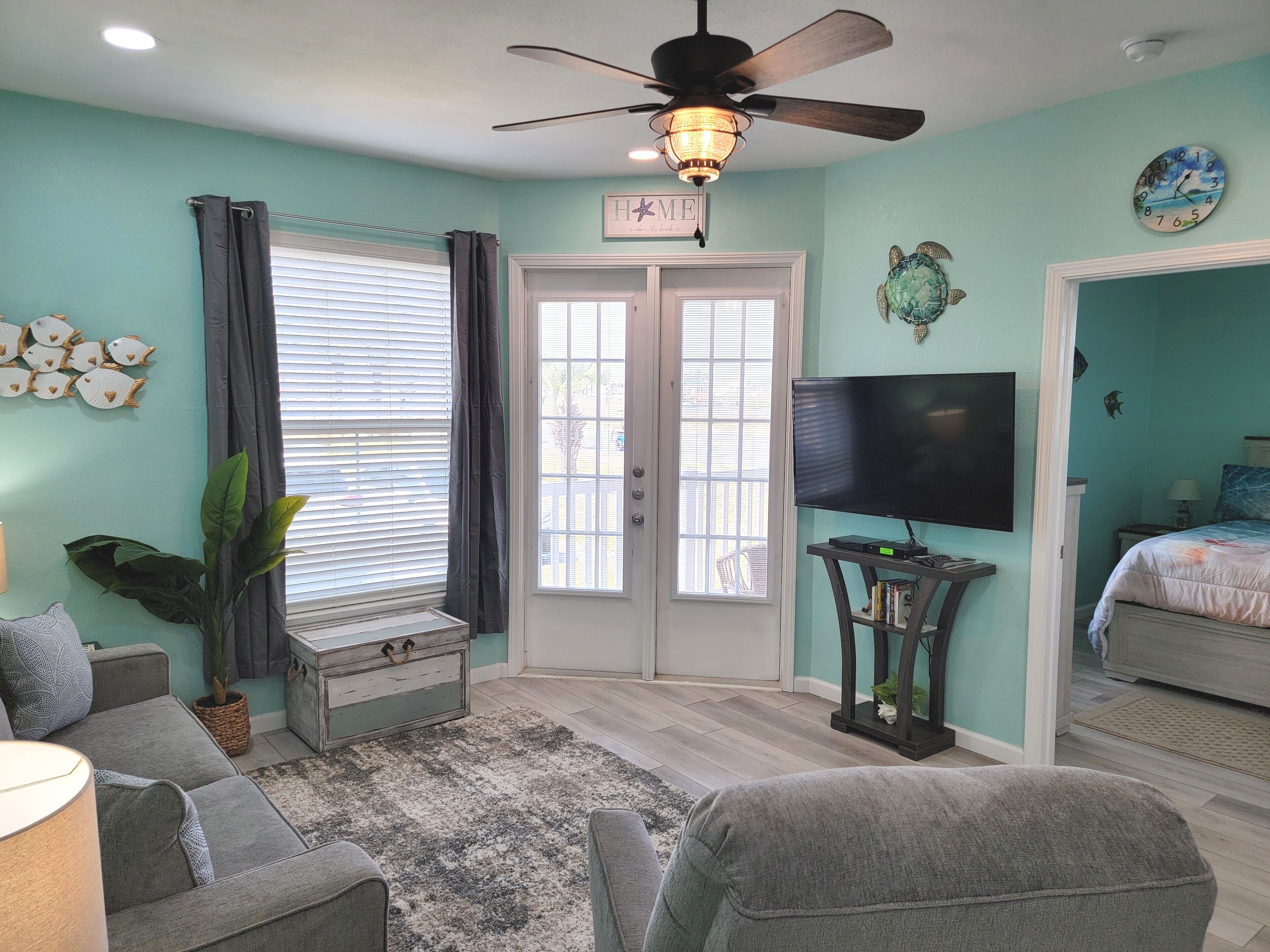 A living room with teal walls, a ceiling fan, a flat-screen TV, a sofa, a coffee table, and a door leading to another room.