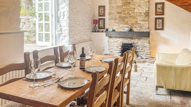 A dining area with a wooden table set for four, surrounded by wooden chairs. A stone fireplace is visible in the background, and a white sofa is positioned to the right. - 3 bedroom accommodation in Swanage (Swanage)