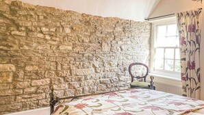 A bedroom with a stone wall, a wooden bed frame, a floral bedspread, and a window with curtains. - 3 bedroom accommodation in Swanage (Swanage)