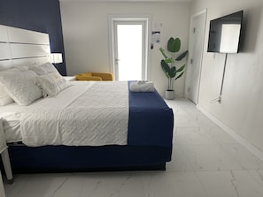 Un lit bien fait, recouvert d’une couvre-lit blanche et bleue, une table de chevet avec une lampe, une plante en pot et un téléviseur à écran plat fixé au mur.