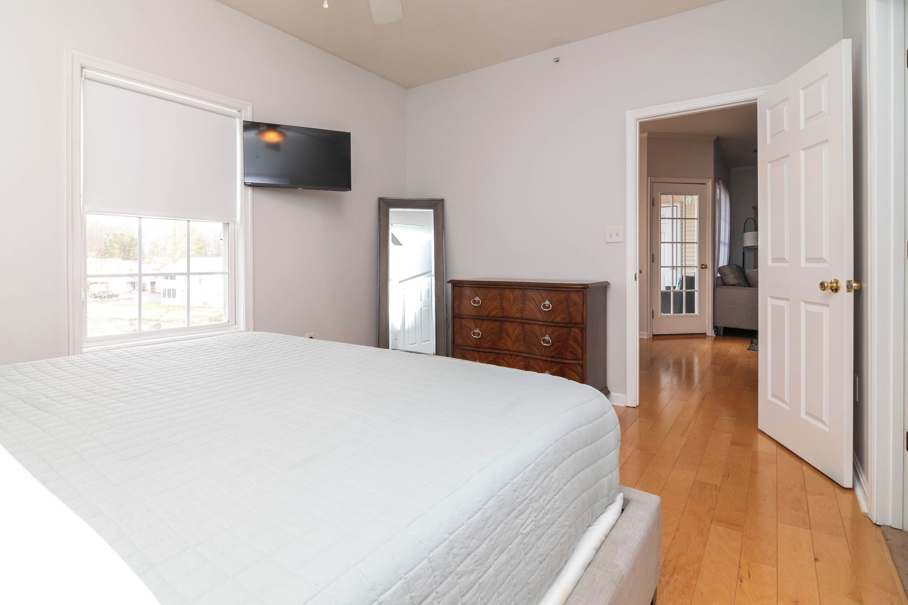 A bedroom with a large bed, a dresser, a TV mounted on the wall, and a door leading to another room.