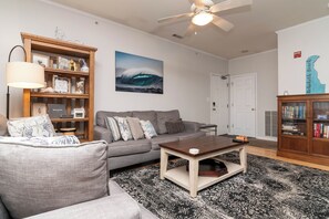 A living room with a grey sofa, a wooden coffee table, a bookshelf, and a ceiling fan.