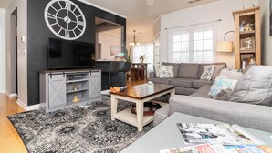 A living room with a large wall clock, a flat-screen TV, a grey sofa, a coffee table, and a bookshelf. -  Cozy Creekwood Getaway on the Highway (Rehoboth Beach)