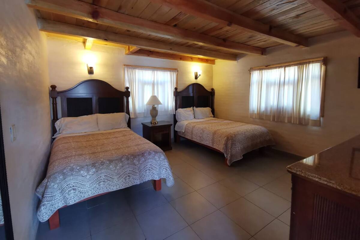 A room with two beds, wooden headboards, a nightstand with a lamp, and a window with curtains.