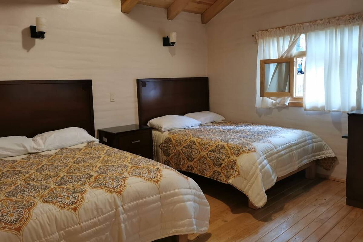 A room with two beds, wooden beams on the ceiling, a window with curtains, and a small table.