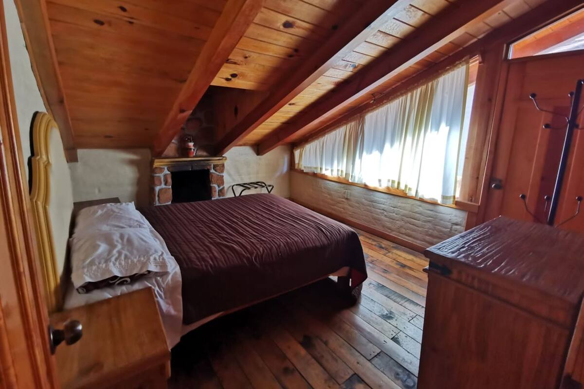 A bedroom with a wooden ceiling, a bed with a brown bedspread, a small table with a lamp, and a window with curtains.