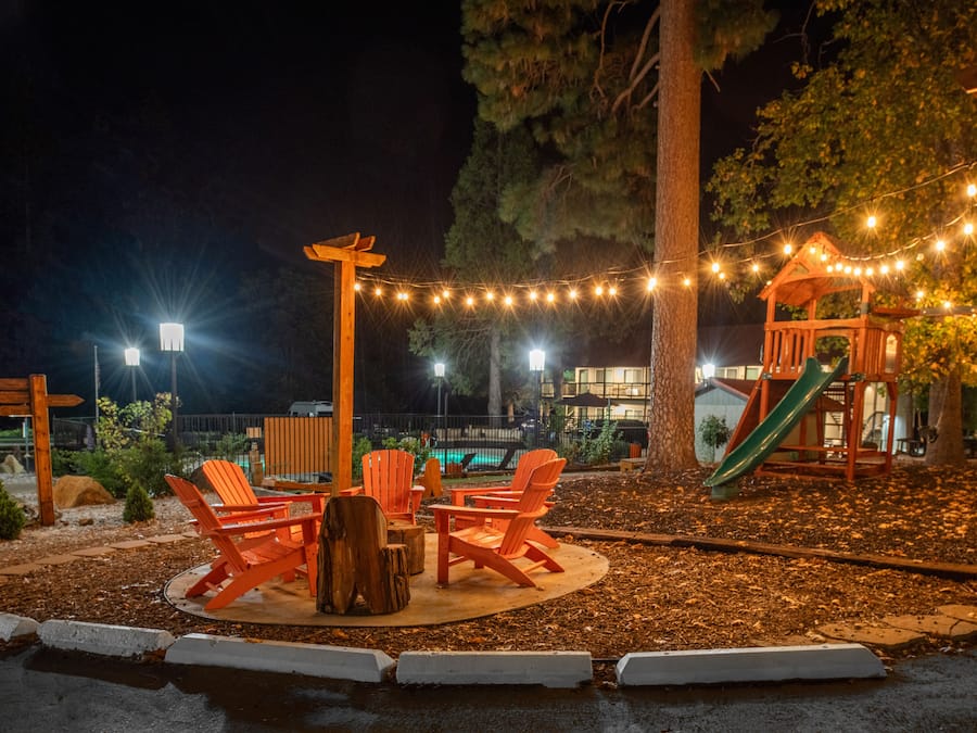 Un espace extérieur aménagé avec des chaises et une table en bois, entouré d’arbres et illuminé de guirlandes lumineuses.