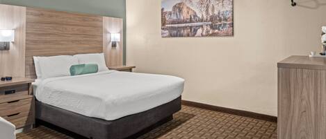 A hotel room with a bed, bedside tables, a nightstand, a desk, a chair, a lamp, a picture on the wall, and a patterned carpet.