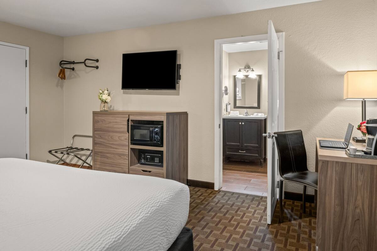A hotel room with a bed, a desk with a computer, a TV, a microwave, and a bathroom visible through an open door.
