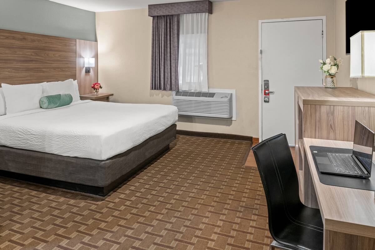 A hotel room with a bed, a desk with a laptop, an air conditioner, a nightstand with a lamp, and a door.