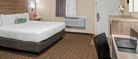 A hotel room with a bed, a desk with a laptop, an air conditioner, a nightstand with a lamp, and a door.