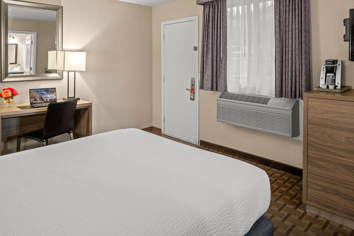 A hotel room with a bed, a desk with a chair, a mirror, a lamp, a door, a window with curtains, and a coffee machine.