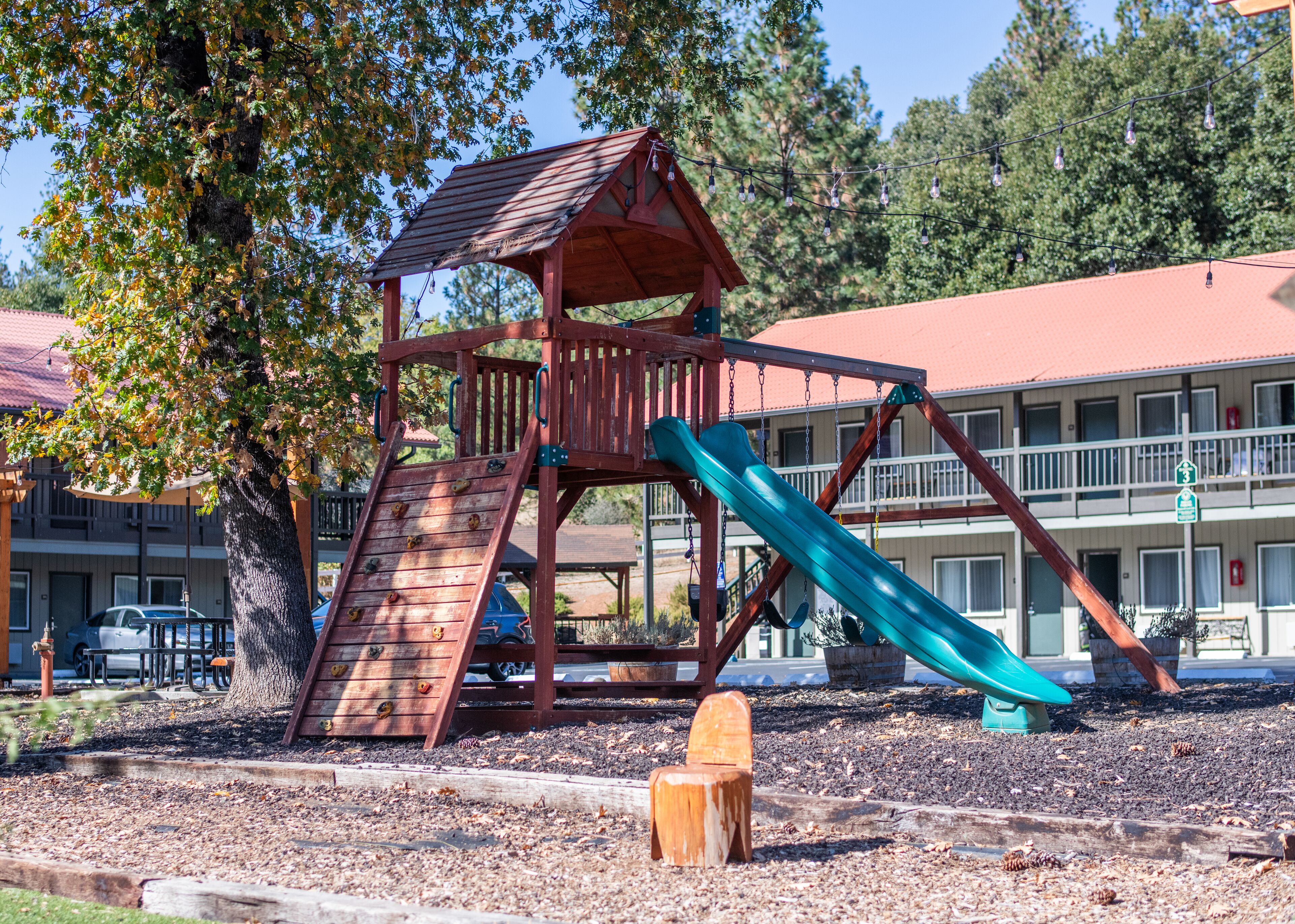 A playground with a slide and climbing area, surrounded by trees and a building with a red roof in the background.