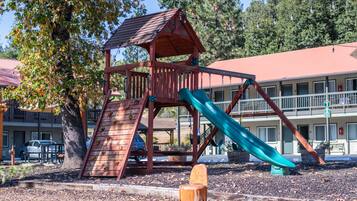 A playground with a slide and climbing area, surrounded by trees and a building with a red roof in the background.