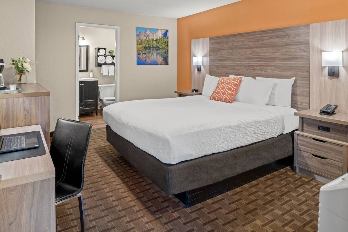 A hotel room with a large bed, a desk with a laptop, a chair, a bathroom with a mirror, and a painting on the wall.