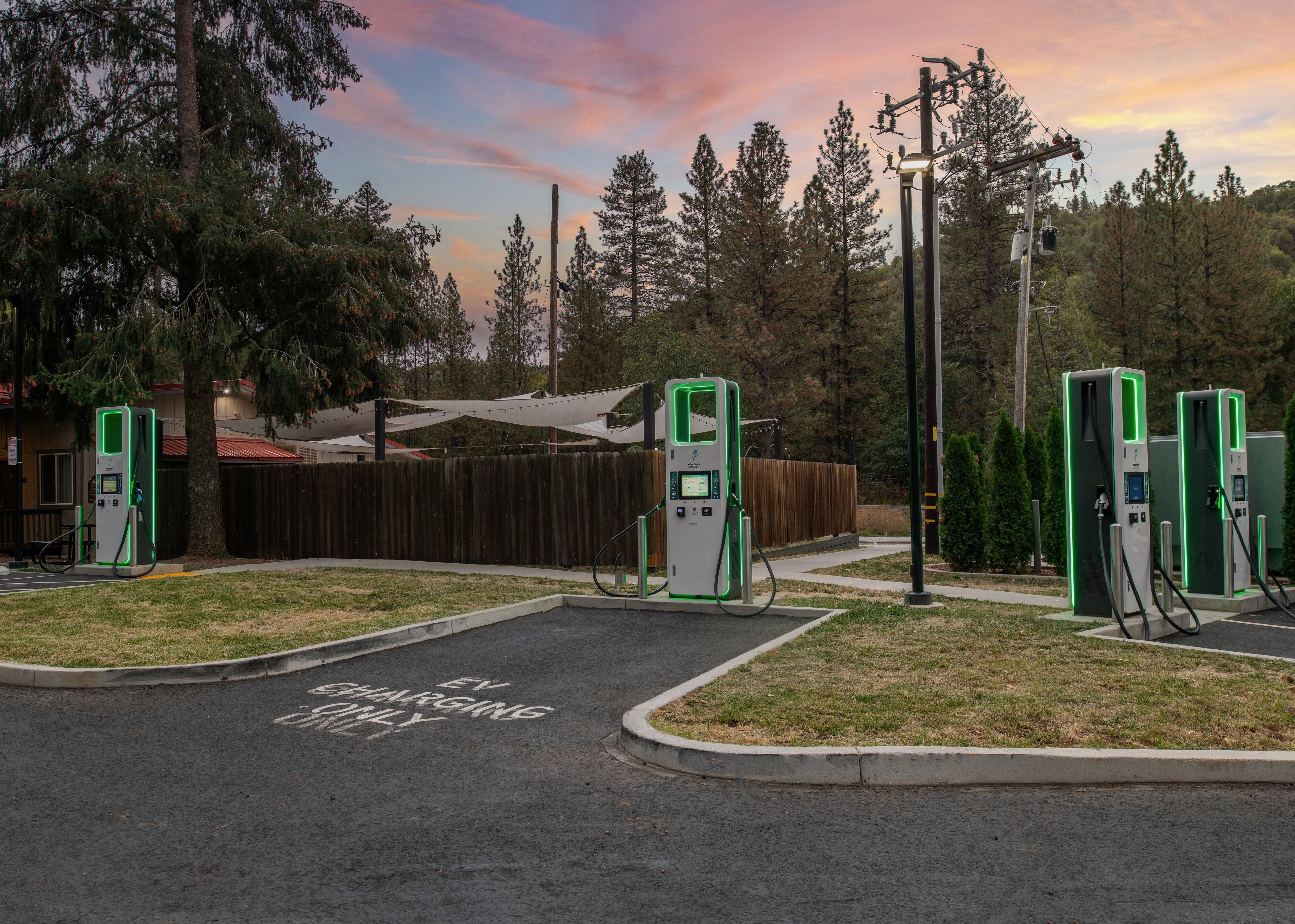 A parking lot with electric vehicle charging stations, a wooden fence, and a building with a red roof in the background.