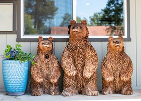 Tres esculturas de madera de osos en un porche con una maceta azul texturizada a la izquierda.