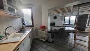 Angolo cottura compatto con forno a microonde, lavello e finestra. C'è un'area pranzo con tavolo e sedie, e un soppalco accessibile tramite una scala in legno.