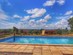 Ein Schwimmbecken mit gefliestem Deck, umgeben von Grünpflanzen und blauem Himmel mit Wolken.