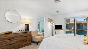A modern bedroom with a wooden dresser, a round mirror, a beige armchair, a large bed with white bedding, a TV on a stand, and a view of a swimming pool through a glass door. - Serene 1 bed Casita w/Pool, Spa & Mountain Views (Palm Springs)