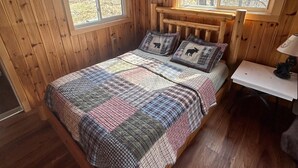 A wooden cabin bedroom with a bed, two windows, and a small table.