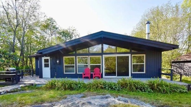 A modern cabin with large windows, a covered porch, and red chairs outside.