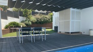 Una piscina con borde de azulejos, rodeada por una terraza de madera y una zona de estar cubierta con sillas y una mesa.
