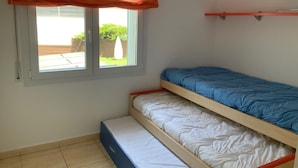Una habitación pequeña con una cama nido, una ventana con cortina translúcida y una estantería sobre la cama.