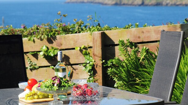 Ein Tisch mit einer Flasche, Salat, Trauben und einem Glas Wasser, vor einer schönen Aussicht auf das Meer und die Hügel.