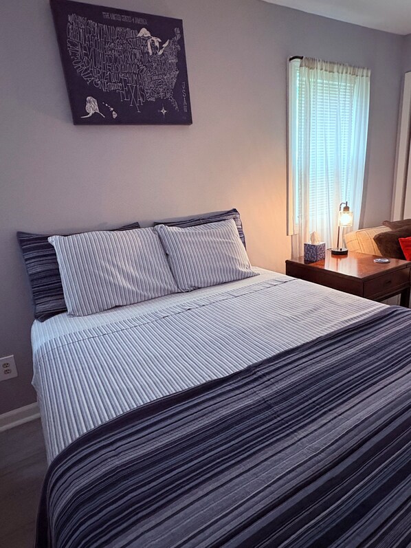A bed with striped bedding, a nightstand with a lamp, a window with curtains, and a wall map.