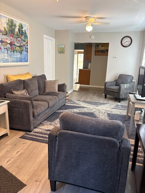 A living room with a grey sofa, a grey armchair, a small table, and a ceiling fan.
