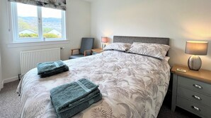 A neatly made bed with a floral pattern, a chair, a bedside table with a lamp, and a window with a view of the outdoors.
