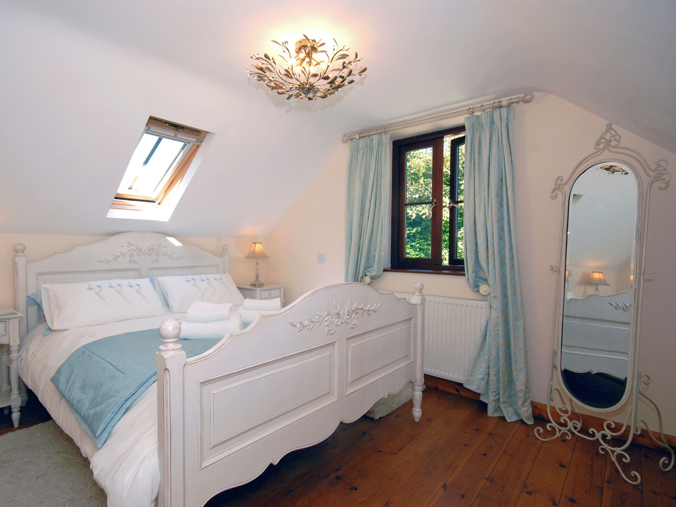 Een slaapkamer met een wit bed, een nachtkastje, een lamp, een raam met gordijnen, een plafondlamp en een spiegel.