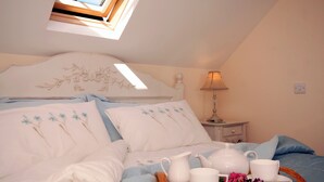 Un dormitorio con tragaluz, cama con ropa de cama floral, mesita de noche con lámpara y bandeja con juego de té y flores.