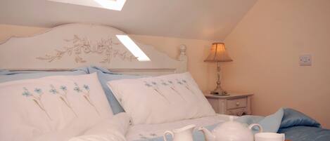 A bedroom with a skylight, a bed with floral bedding, a bedside table with a lamp, and a tray with tea set and flowers.