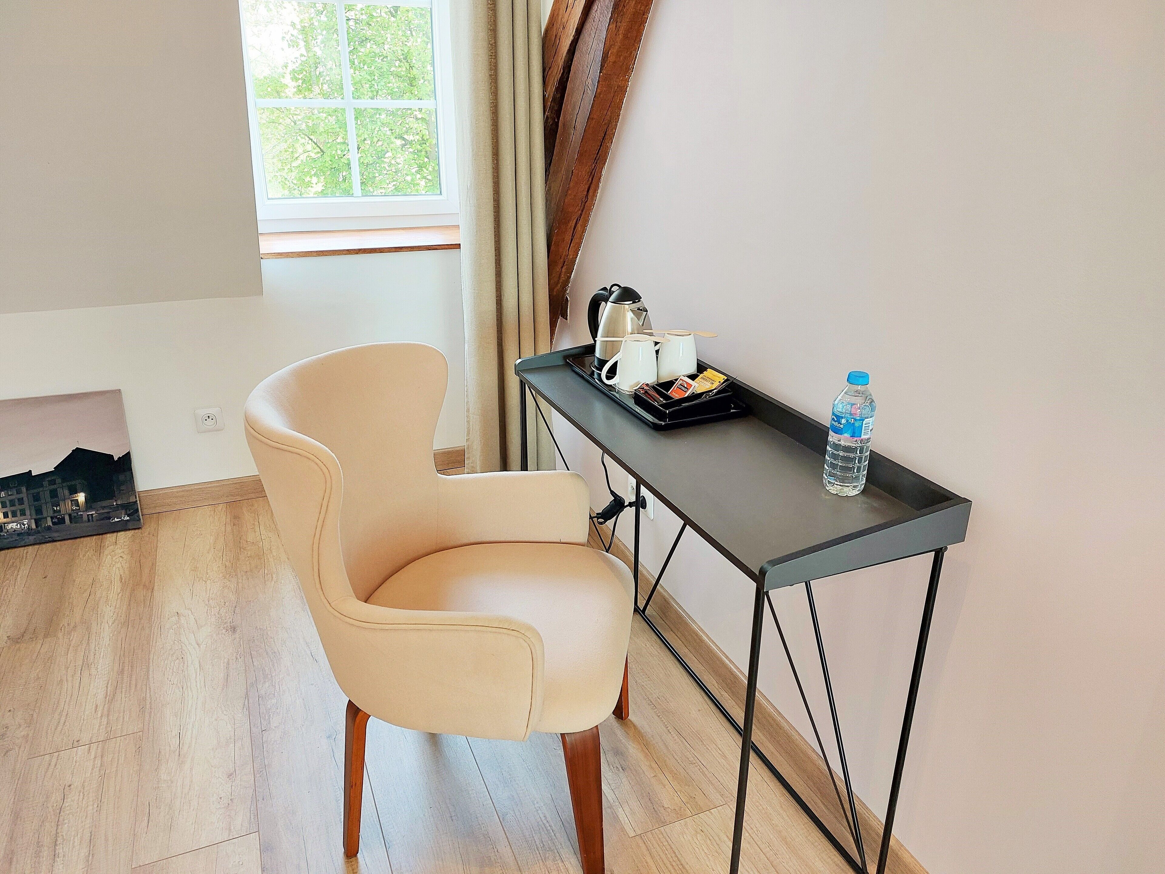 Une pièce avec un fauteuil beige, une petite table sur laquelle se trouve une bouteille d’eau, un plateau avec des tasses et une bouilloire, et une fenêtre avec des rideaux.