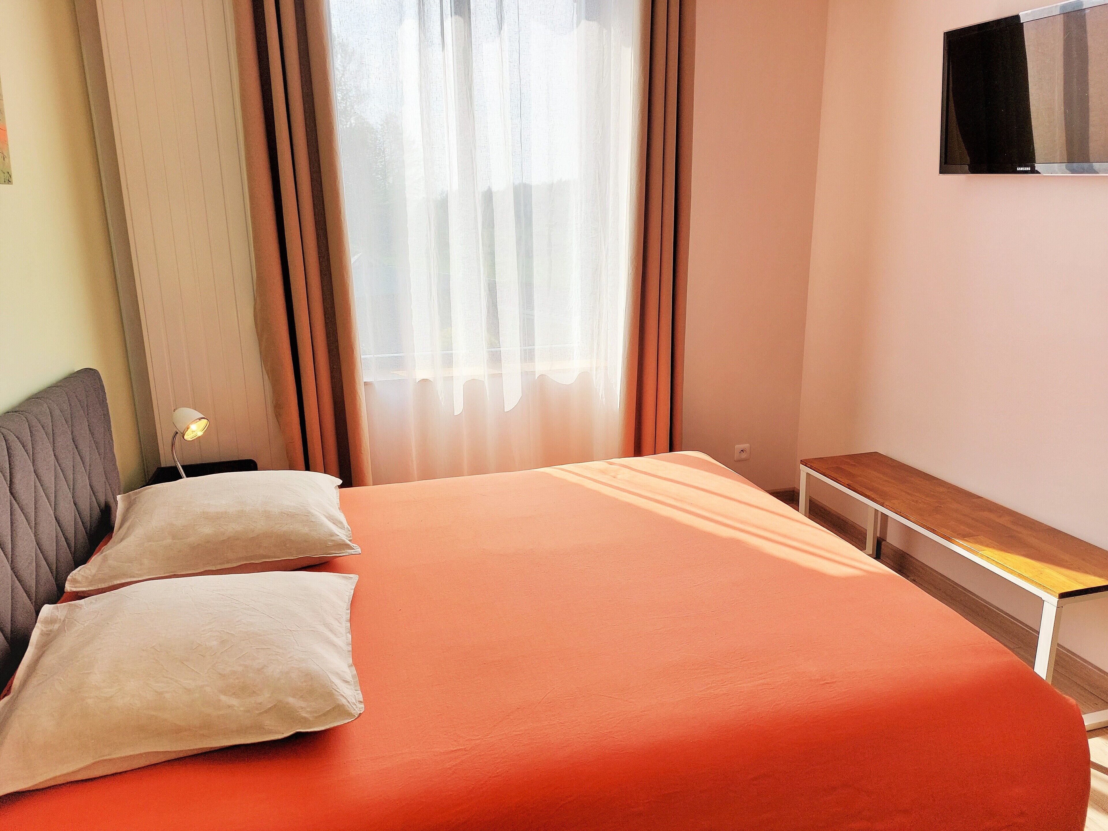 A bedroom with a bed, pillows, a bedside table, a lamp, a TV mounted on the wall, and a window with curtains.