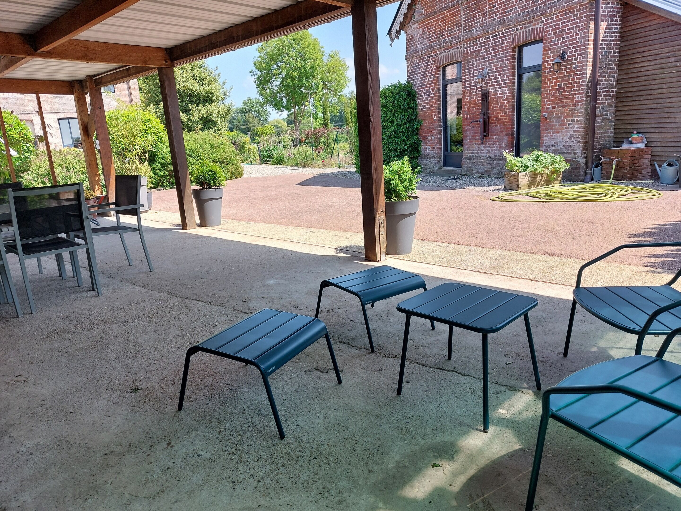 Un espace extérieur abrité avec des tables et des chaises en métal, un bâtiment en briques en arrière-plan et des plantes en pot.