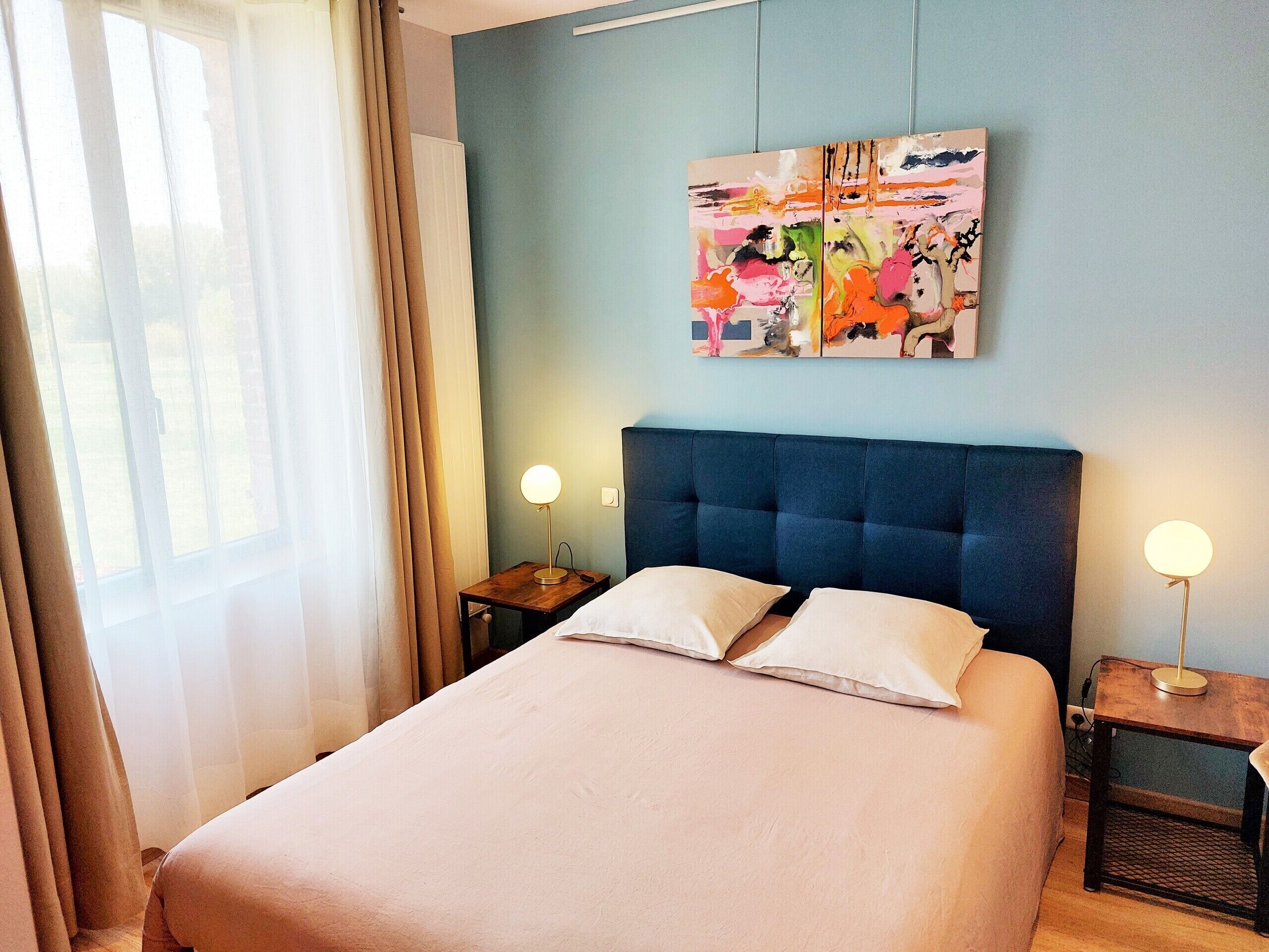 A hotel room with a large bed, two bedside tables, a wall-mounted painting, and a window with curtains.