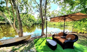 A serene outdoor seating area with a wicker sofa and an umbrella, overlooking a calm body of water and lush greenery.