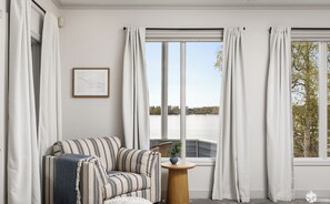 A cozy room with a striped sofa, a small round table with a plant, and a view of a lake through large windows with white curtains.