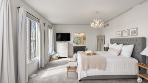 A bedroom with a large bed, a TV, a vanity with a mirror, and a chandelier.