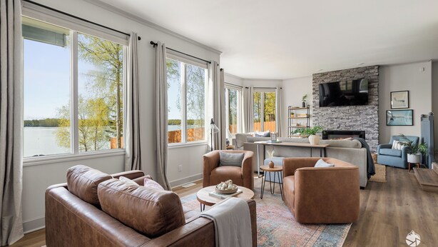 A spacious living room with a leather sofa, a coffee table, and a fireplace. There are large windows with curtains, a stone fireplace wall, and a television mounted above it.