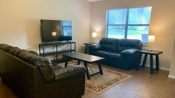 Una sala de estar con un sofá, una mesa de centro, un televisor sobre un mueble bajo y una ventana con persianas.