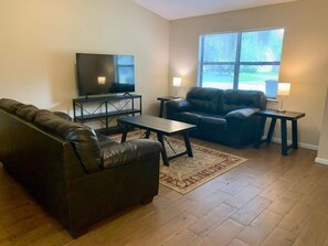 A living room with a sofa, a coffee table, a TV on a stand, and a window with blinds. - Wonderful 3 Bedroom 2 Bath (St. Marys)