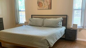 Un dormitorio con cama, ventilador de techo, un cuadro en la pared y ventanas con cortinas.
