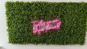 Un canapé gris agrémenté de deux coussins roses, placé contre un mur dont le fond est vert et texturé, et sur lequel on peut voir un panneau néon avec l’inscription : « Ensemble, nous pouvons conquérir le monde. »