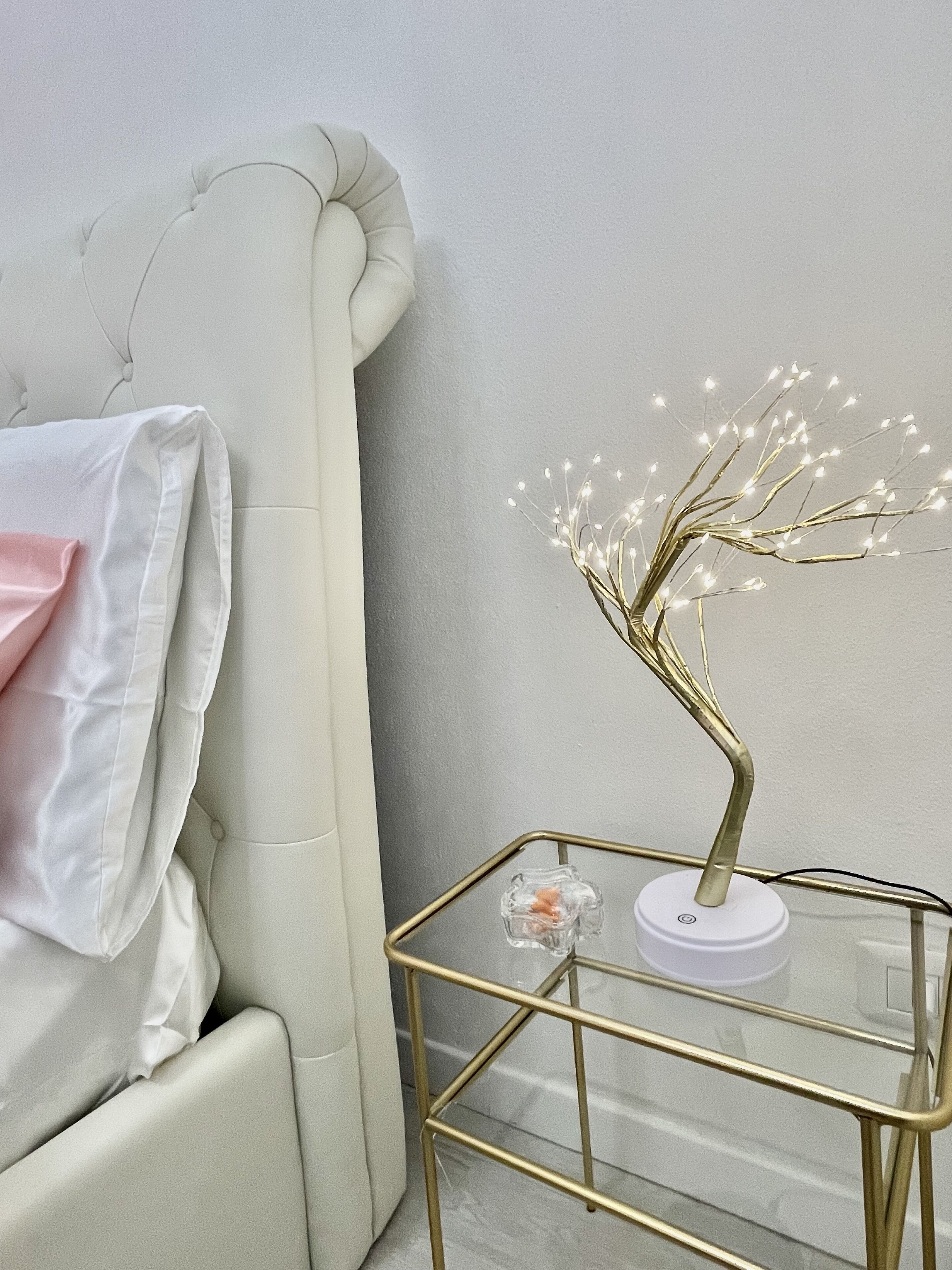Ein Zimmer mit einem weißen, gepolsterten Kopfteil, einem Glastisch mit goldener Rahmen und einem geschmückten Baum mit Lichtern.