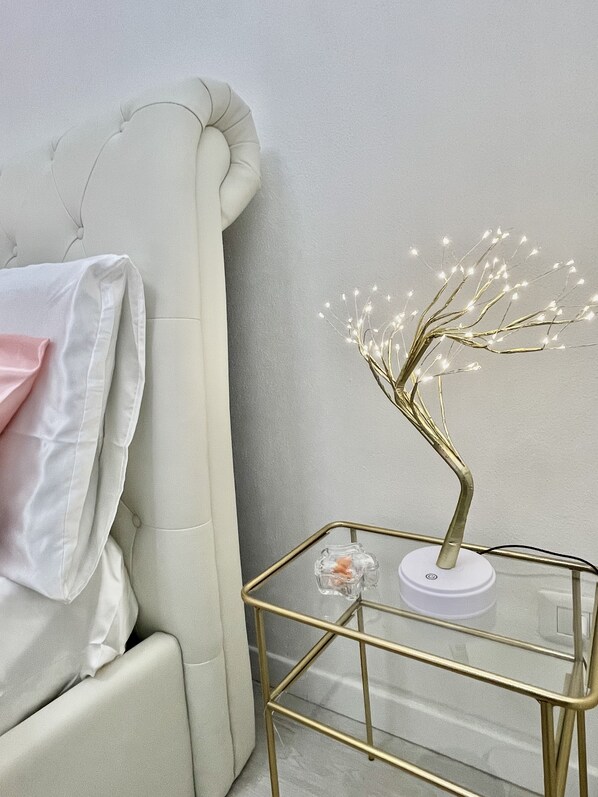 A room with a white upholstered headboard, a glass coffee table with a gold frame, and a decorative tree with lights.