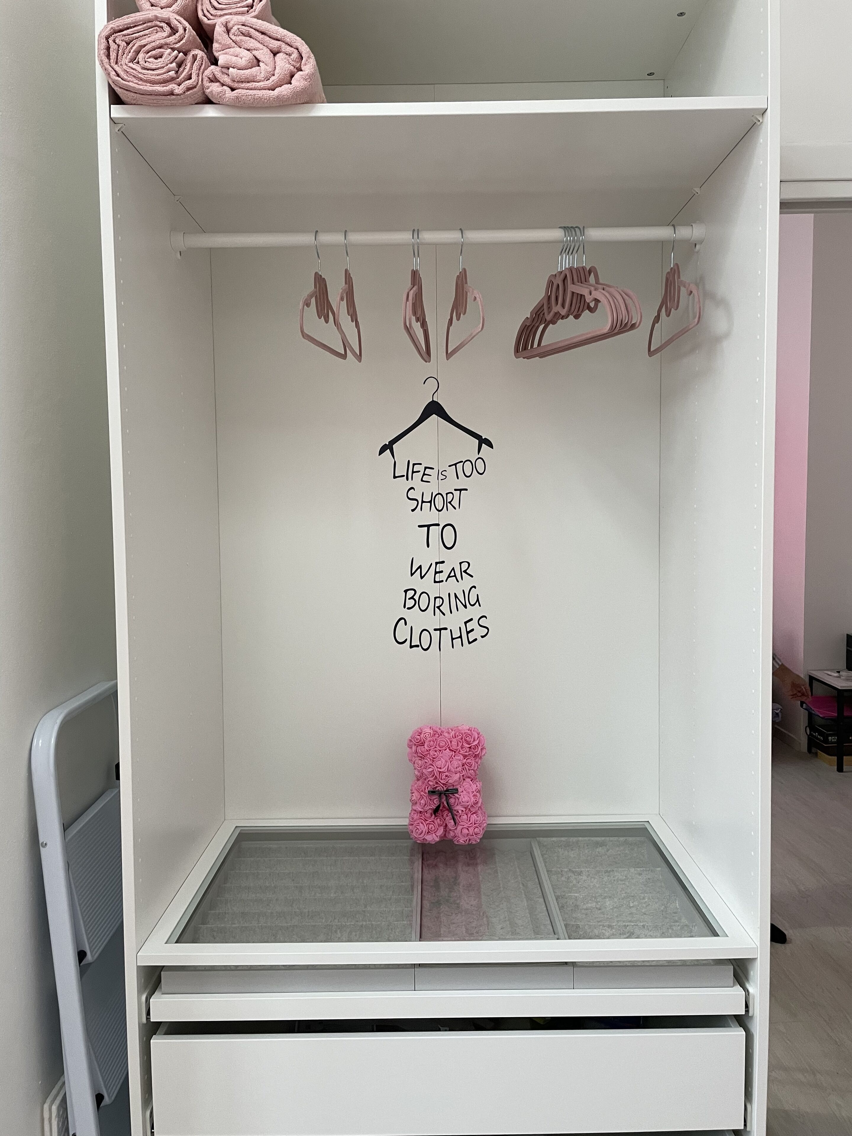 A white wardrobe with a mirror, a hanging rod with clothes, and a motivational quote on the wall.